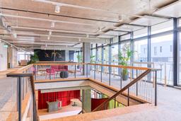 Open, modern coworking space at iCITY ALBA HAUS with a view of the guest reception area. Visible are large windows, vibrant lounge furniture, and a wooden staircase leading to the reception below, designed for a welcoming and professional atmosphere. Offener, moderner Coworking-Bereich im iCITY ALBA HAUS mit Blick auf den Gästebereich. Sichtbar sind große Fenster, lebhafte Loungemöbel und eine Holztreppe, die zur Rezeption darunter führt, gestaltet für eine einladende und professionelle Atmosphäre.